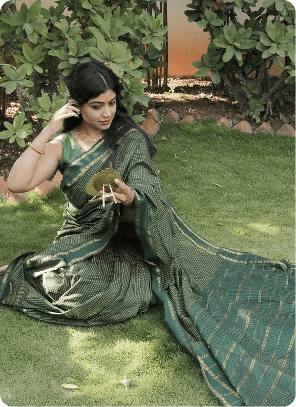 Woman in a green saree sitting on grass with plants in the background